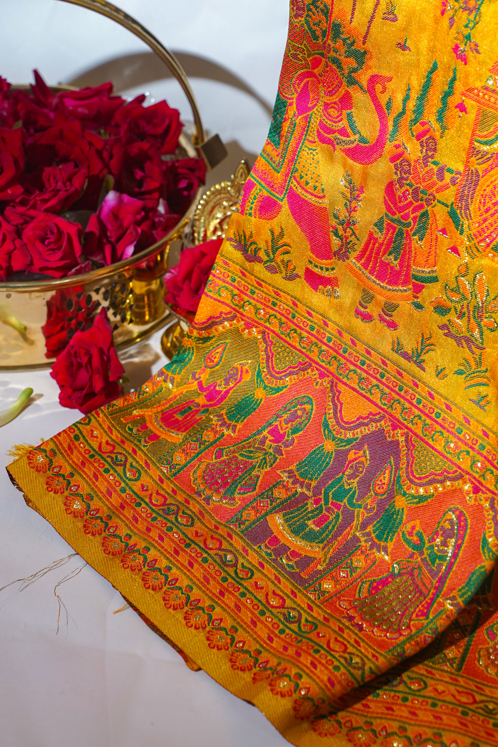 Decorative fabric with intricate patterns and colors, placed on a white surface with red flowers in the background.