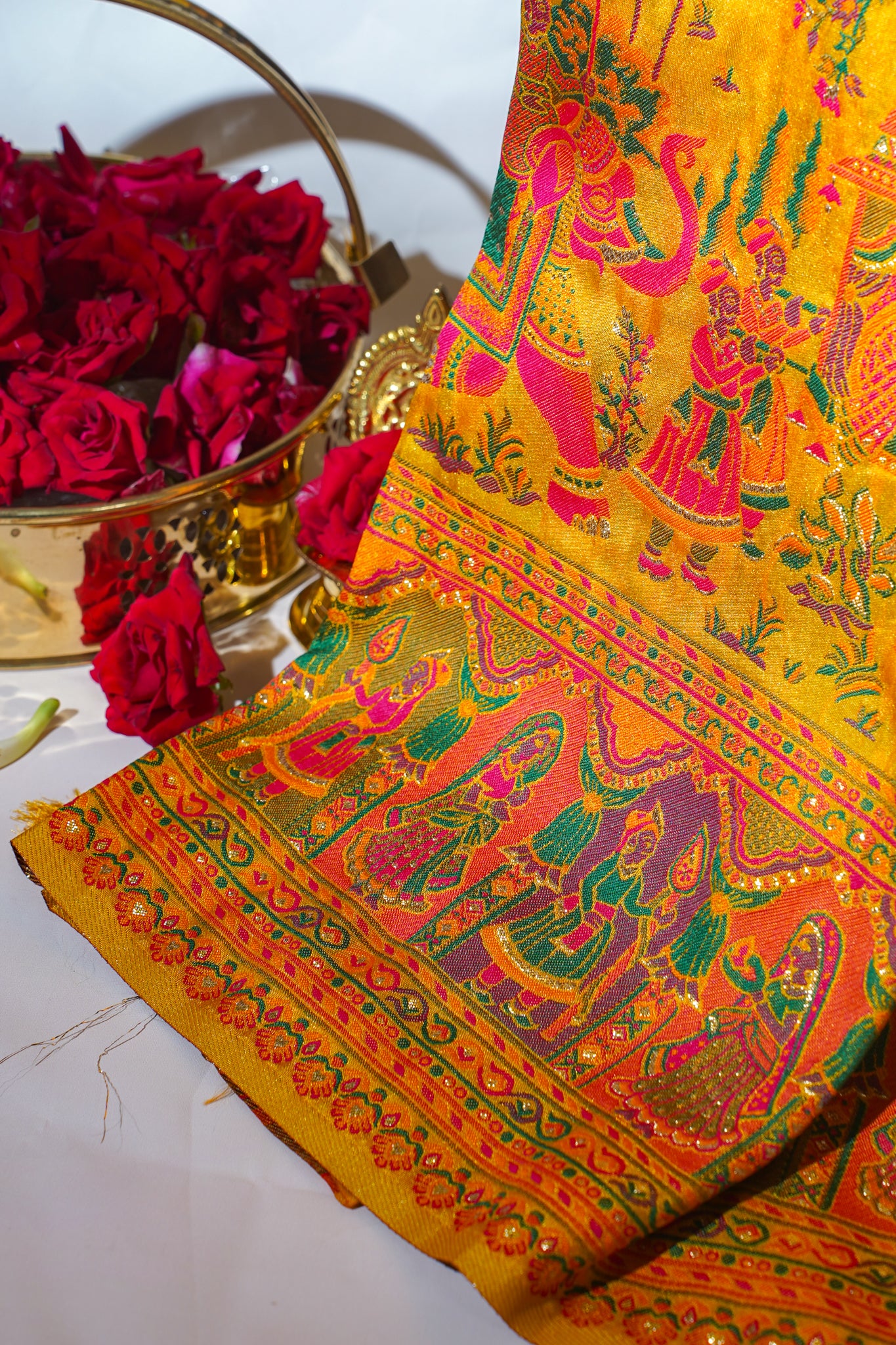 Decorative fabric with intricate patterns and colors, placed on a white surface with red flowers in the background.