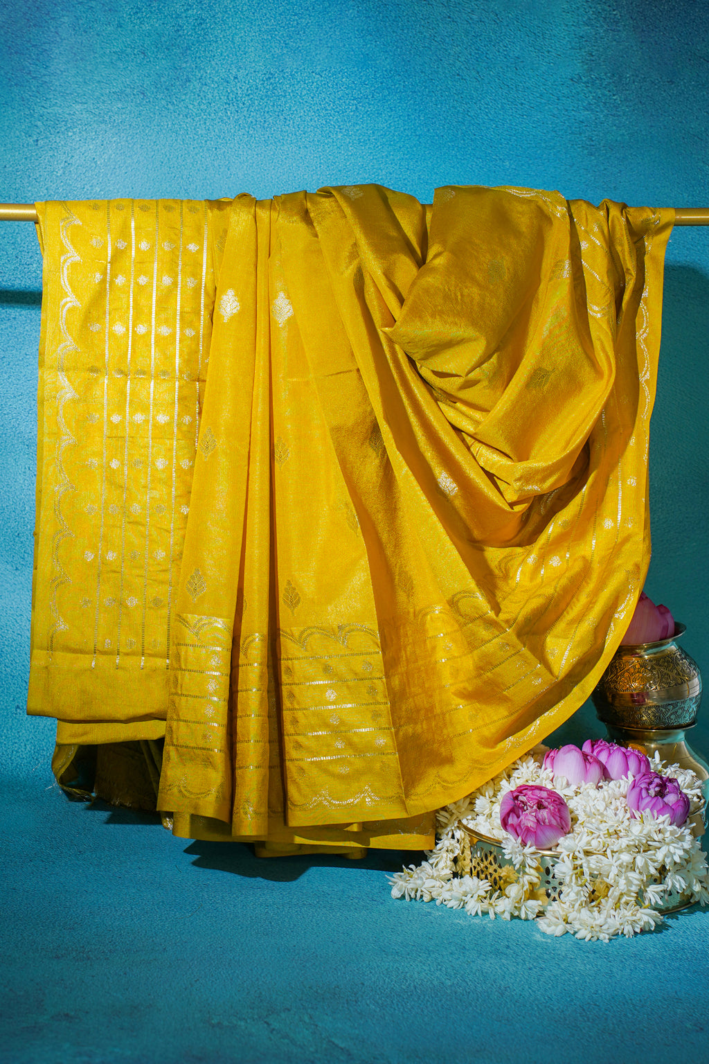 Yellow saree draped over a rod with flowers and a pot on a blue background