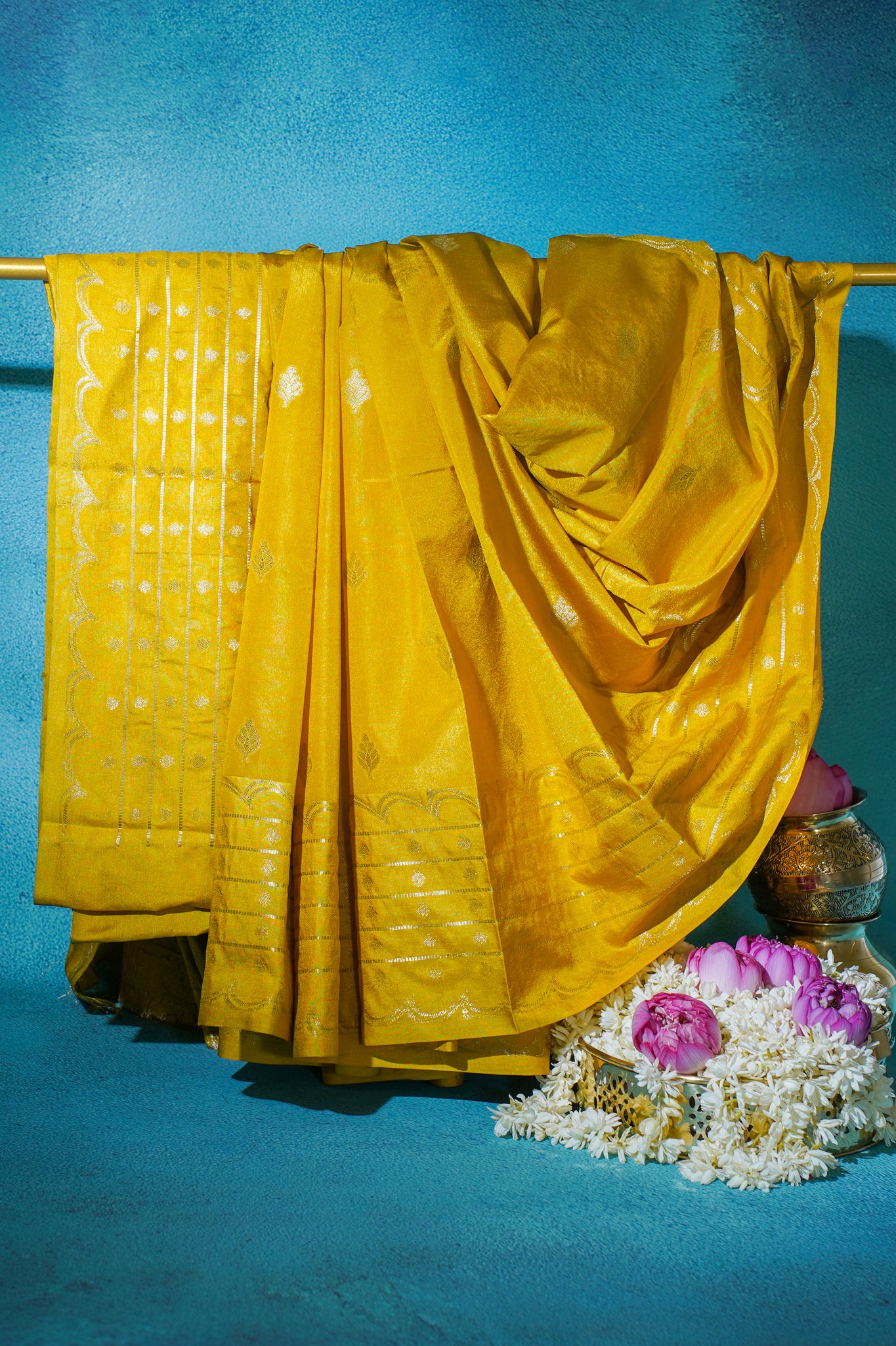 Yellow saree draped over a rod with flowers and a pot on a blue background